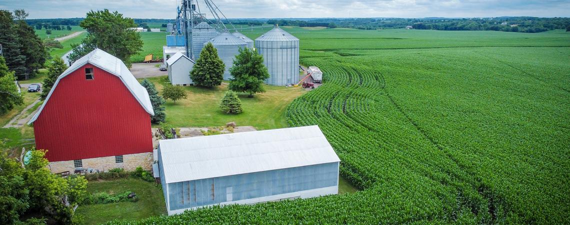 Aerial view of red barn and metal grain bins standing next to green corn field.