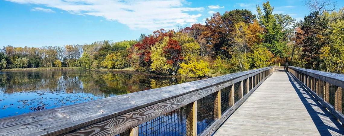 A wooden walkway over a small lake surrounded by red, yellow, and green trees.