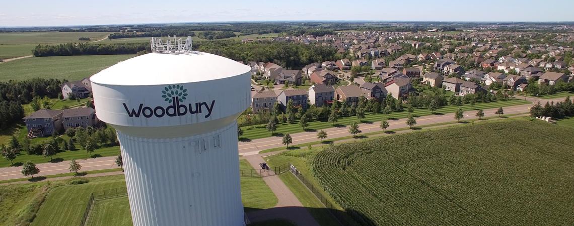 A white water tower labeled Woodbury stands next to a corn field with a suburban neighborhood in the background.
